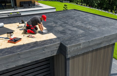 A construction worker applies EPDM roofing material on a flat roof of a modern home in a sunny outdoor environment