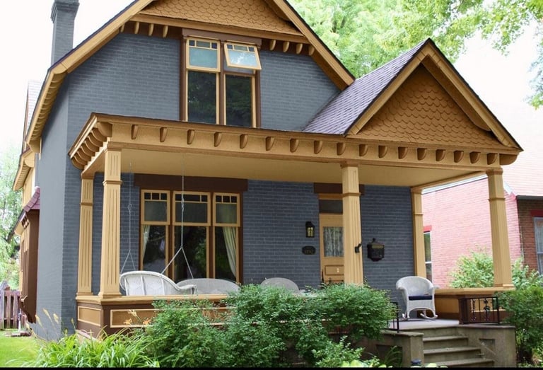 Blue and tan Victorian-style house with wooden front porch, multiple gables, and landscaped front lawn