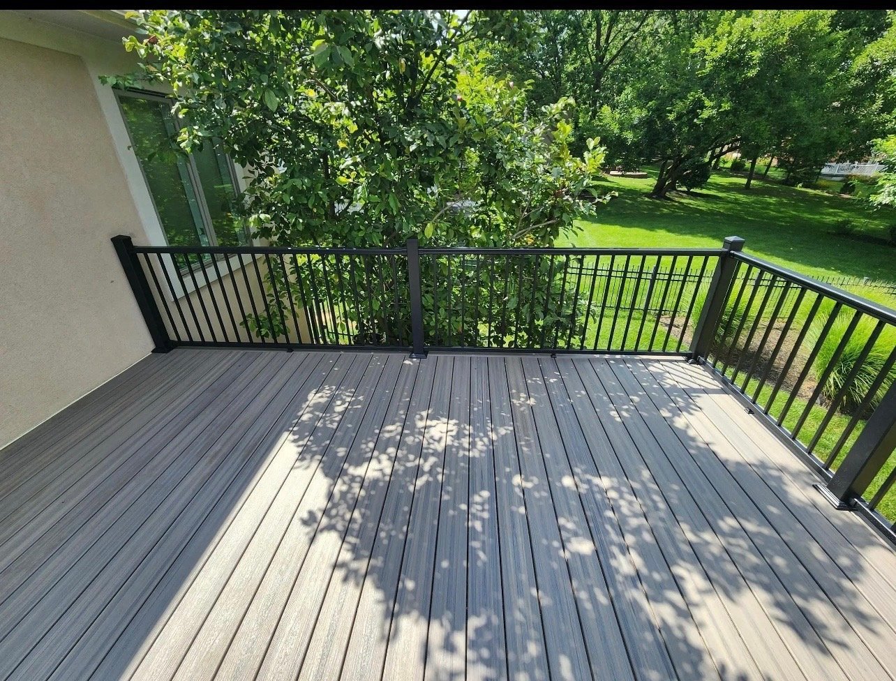 A spacious gray composite deck with black railings overlooking a green lawn and tree-lined yard on a sunny day