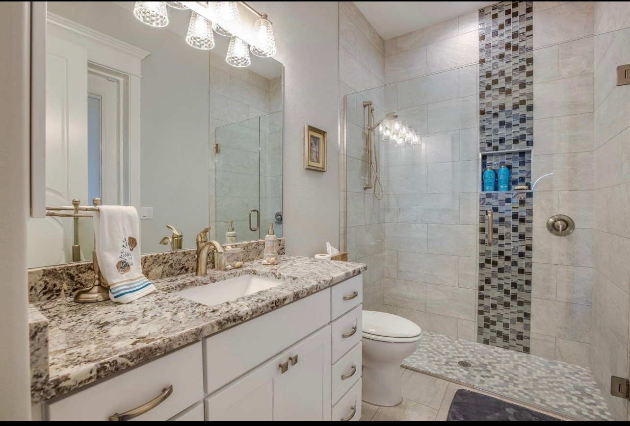 Modern bathroom with white vanity, granite countertop, large mirror with lighting, and walk-in shower with tile accents