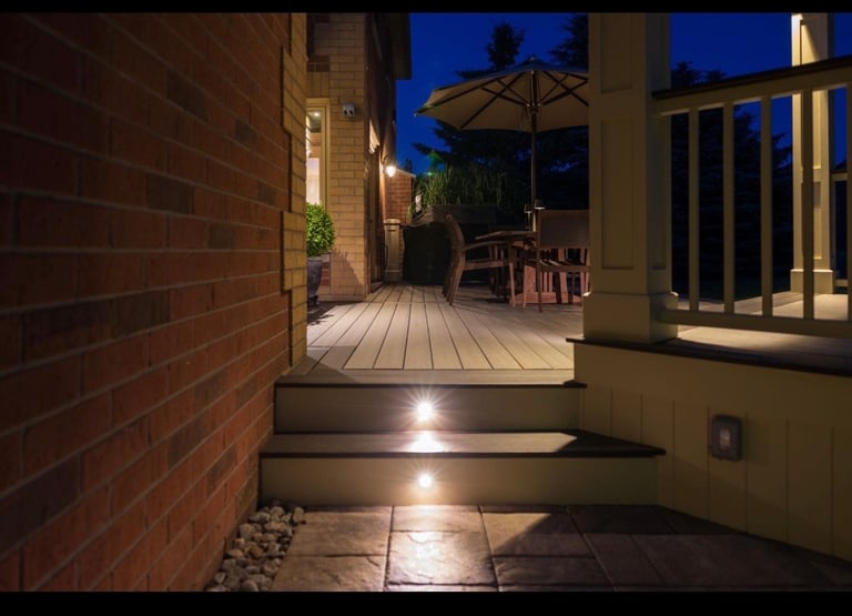Illuminated outdoor patio and deck at dusk with brick wall, white columns, umbrella, and string lights