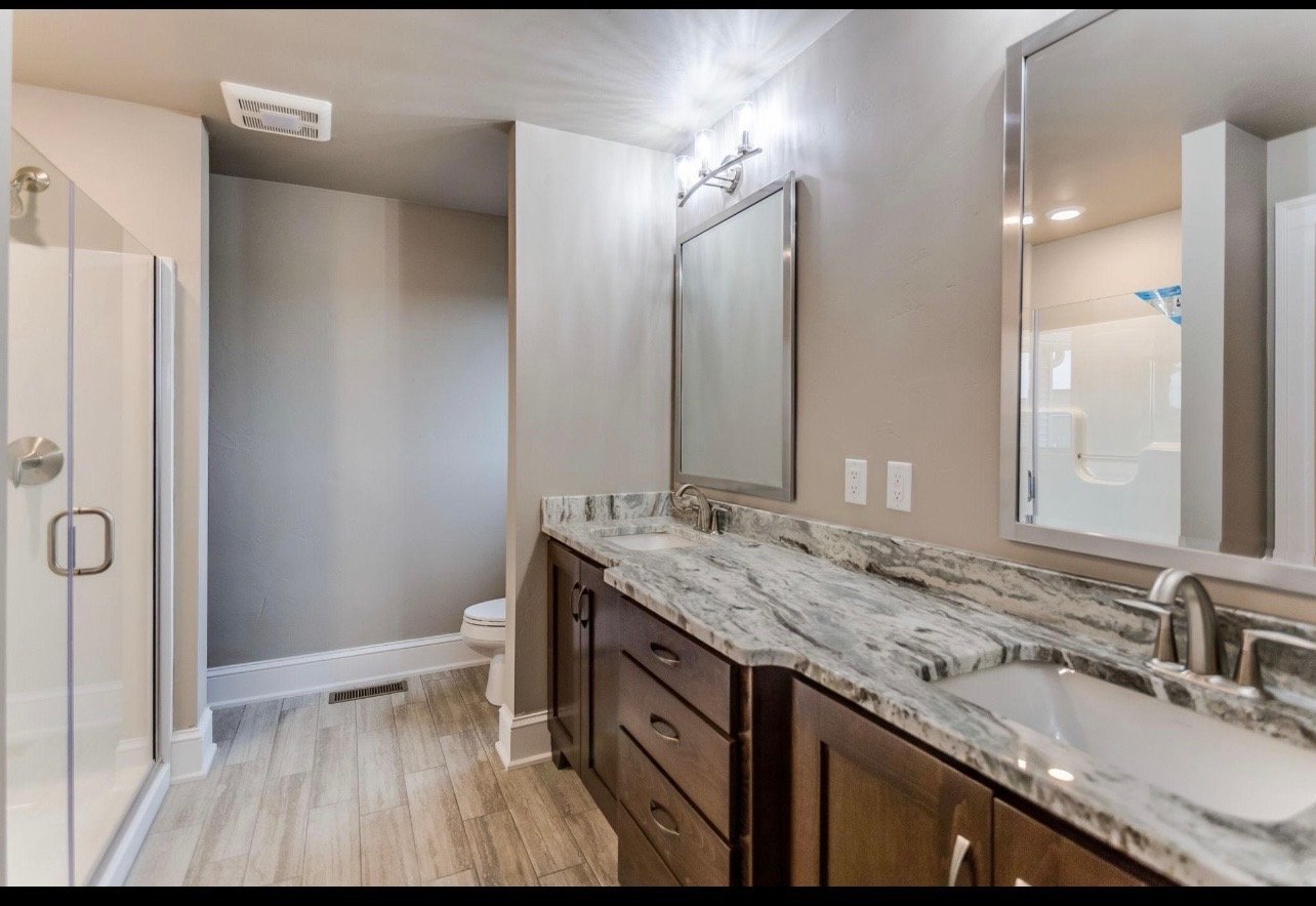 Modern master bathroom with dark vanity, marble countertops, dual sinks, and glass-enclosed shower area