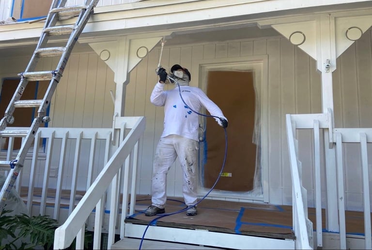 Painter in white coveralls spraying exterior house porch with paint gun on front entrance