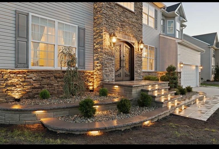 Modern home exterior at dusk with illuminated stone accents, landscaping lights, and decorative shrubs on the front entryway
