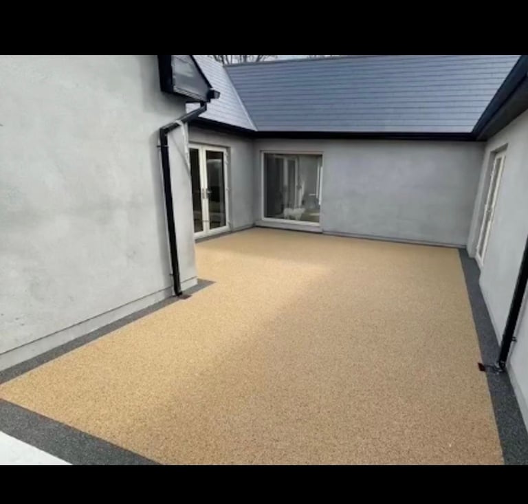 Modern patio area with tan permeable paving, white-painted walls, dark roof, and black-framed windows and doors